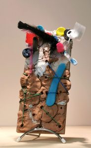 Masque. H = 30 cm. Sur planche à trous. Argile, objets de recyclage. Liens, plumes.